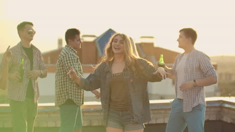 The-beautiful-American-girl-is-dancing-on-the-roof-with-her-five-friends-on-the-party-who-drinks-beer-from-green-bottles.-She-smiles-and-enjoys-the-time-in-shorts-and-a-light-denim-jacket-in-summer-evening.-Her-blond-hair-is-flying-in-the-wind.